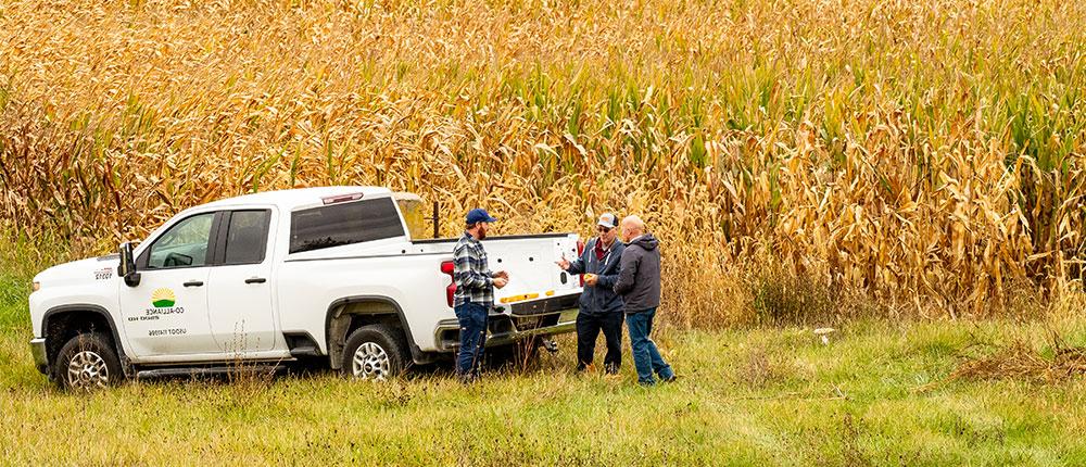 Three men talking by a truck with a Co-Alliance logo on the side, with a corn field in the background